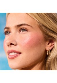 Nærbilde av en persons ansikt med lyst blont hår, klar hud, subtile rosa kinn og gullhengerør med minimalistisk design.