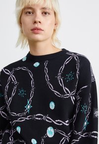 Sweater preto com um design padronizado, apresentando formas em roxo claro e turquesa, incluindo círculos e estrelas, com gola e punhos canelados.