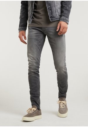 Mann trägt verblasste graue Skinny-Jeans, graue Sneaker mit cremefarbenen Schnürsenkeln, ein dunkelgraues T-Shirt und eine gewaschene schwarze Jeansjacke.