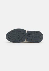 Semelle de chaussure de sport en caoutchouc noir avec des motifs texturés, présentant des rainures et une bande d'accent beige. Nom de la marque embossé sur la semelle.