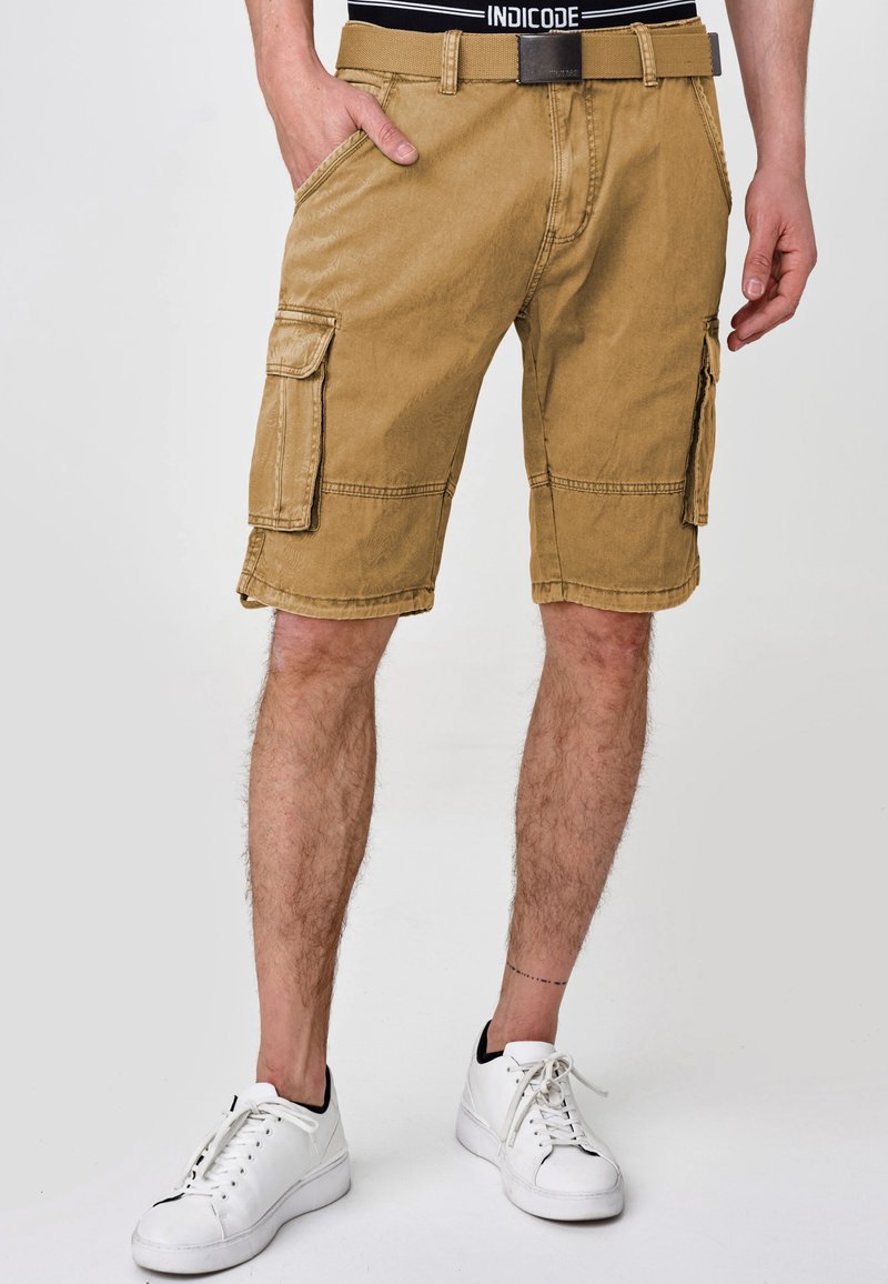 Khaki cargo shorts dengan potongan santai, beberapa kantong samping, dan detail jahitan. Dipadukan dengan sepatu sneakers putih dan ikat pinggang hitam.