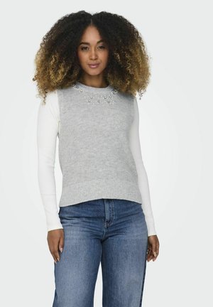 Grauer Strickpullover mit einem mit Perlen verzierten Ausschnitt, übereinander getragen mit einem weißen Langarmoberteil. Kombiniert mit blauen Denim-Jeans mit geradem Schnitt.