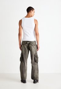 Homme portant un débardeur blanc, un pantalon cargo vert olive délavé avec des poches latérales et des bottes noires, se tenant de dos contre un fond blanc.