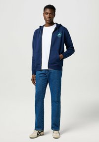 Felpa con zip navy, con tasche frontali e design del logo, abbinata a jeans azzurri chiari e sneakers crema, in piedi contro uno sfondo neutro.