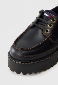 Černé kožené lodní boty s hnědým prošitím, zlatými očky a robustní černou podrážkou. Obsahují decentní logo štítek na jazyku.