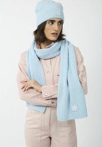 Bonnet tricoté bleu clair et écharpe assortie avec une texture côtelée. L'écharpe drape autour du cou ; les deux présentent un patch logo.