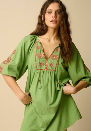 Femme aux longs cheveux ondulés porte une tunique verte ample avec une broderie circulaire rouge et beige et des liens à pompons, debout devant un fond beige.