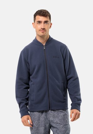 Giacca in pile blu navy con zip e colletto alto, due tasche laterali e una texture sottile. Indossata sopra pantaloni grigi a fantasia.