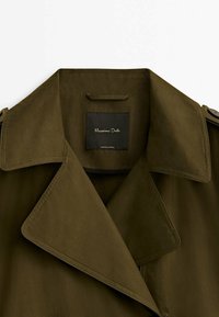 Zielonooliwkowy płaszcz trencher z szerokim kołnierzem, detalami guzików na ramionach oraz czarną metką wewnętrzną z nazwą marki.