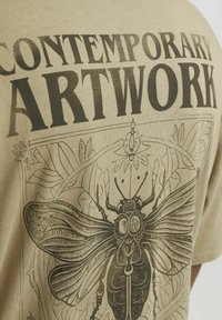 Camiseta de algodón beige con estampado gráfico negro de un escarabajo rodeado de follaje y las palabras "OBRA DE ARTE CONTEMPORÁNEA."