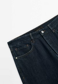 Jean en denim bleu marine avec poche avant, passants pour ceinture, fermeture par bouton métallique et coutures orange visibles, posé à plat sur un fond blanc.