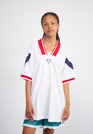 Jersey blanc surdimensionné avec des accents rouges et marins, manches courtes, design en v, arborant un logo au centre. Porté avec un short vert.