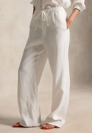 Άτομο που φοράει άνετο, λευκό λινό παντελόνι με κορδόνι και λευκές σανδάλες, στέκεται με το ένα χέρι στην τσέπη μπροστά σε ουδέτερο φόντο.