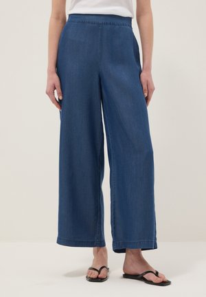 Femme portant un pantalon en jean bleu taille haute à jambes larges, associé à des sandales noires et un top blanc, debout devant un fond uni.