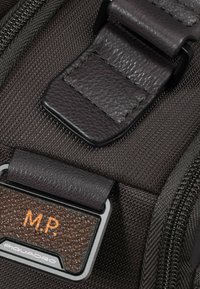 Cinturino in pelle marrone con ferramenta argentata testurizzata, attaccato a uno sfondo di tessuto scuro. Presenta le iniziali "M.P." in rilievo sull'etichetta.