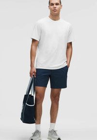 Balts īsām piedurknēm T-krekls, tumši zili teksturēti šorti, pelēki sporta apavi un tumši zilā rokassoma ar baltu akcentu un logo.