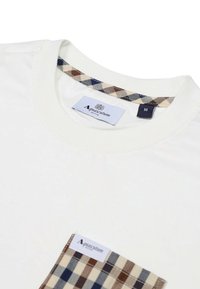 T-shirt bianco in cotone con scollatura rotonda. Presenta una tasca in tessuto a scacchi nei colori marrone, beige e blu, e un'etichetta con la scritta "Aquascutum."