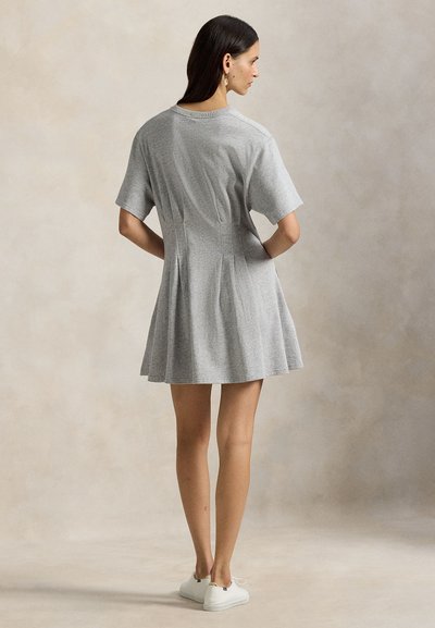 Γυναίκα που στέκεται φορώντας γκρι φόρεμα A-line με κοντά μανίκια και λευκά αθλητικά παπούτσια, με τα χέρια στη μέση, γυρισμένη προς τα πίσω μπροστά σε μπεζ φόντο.