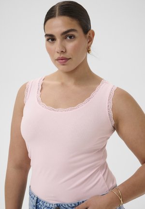 Femme aux cheveux foncés portant un haut rose sans manches avec bordure en dentelle, portant des boucles d'oreilles créoles en or et des bracelets, posant devant un fond uni.