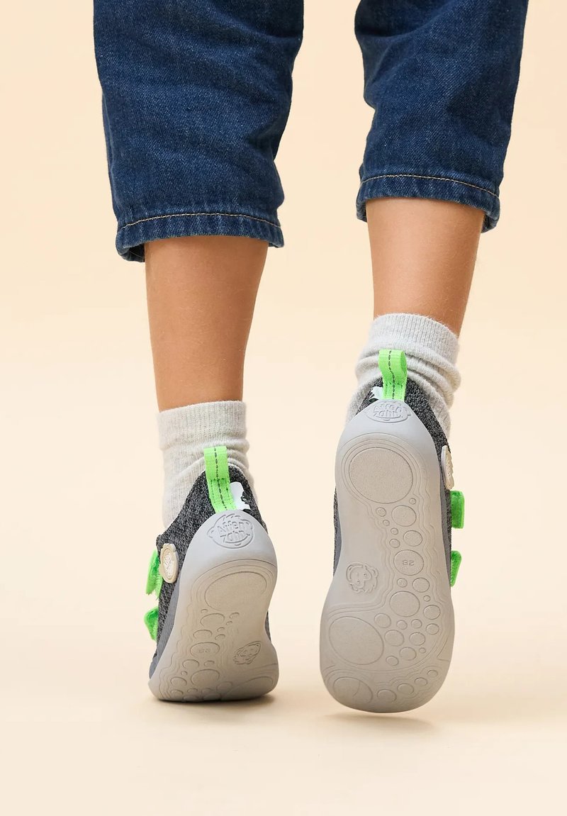 Zapatos de niño con parte superior de textil gris, detalles en verde y suela de goma gris texturizada. Correas ajustables y calcetines grises suaves visibles.