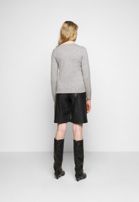 Femme aux cheveux blonds portant un haut gris à manches longues, un short en cuir noir et des bottes noires montantes, debout de dos.