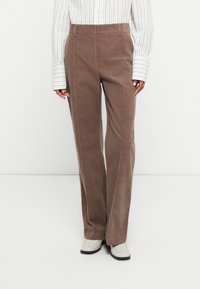 Bruine corduroy broek met voornaad, gedragen met een wit verticaal gestreept shirt en witte vierkante schoenen op een effen achtergrond.