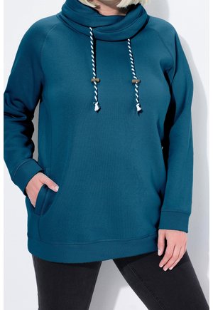 Femme portant un sweat à capuche turquoise avec des cordons blancs et bleus et un pantalon noir, une main dans la poche, debout devant un fond blanc.