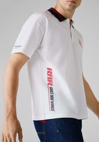 Hvid polo shirt med en marineblå krave, der har rød branding og tekst på siden. Korte ærmer og en slim fit design.