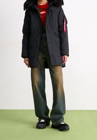 Svart parkas med en huva som är fodrad med päls, har flera fickor och en dragkedja. Kombinerad med urtvättade, vida jeans och kraftiga svarta skor.