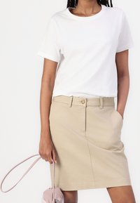 Weißes Baumwoll-T-Shirt, kombiniert mit einem beigen knielangen Rock mit vorderem Knopf, seitlichen Taschen und glatter Textur. Hält eine rosa Handtasche.