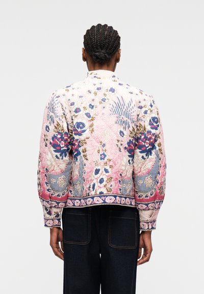 Personne aux cheveux tressés portant une veste à manches longues à imprimé floral avec des motifs roses, bleus et blancs, accompagnée d'un pantalon foncé.