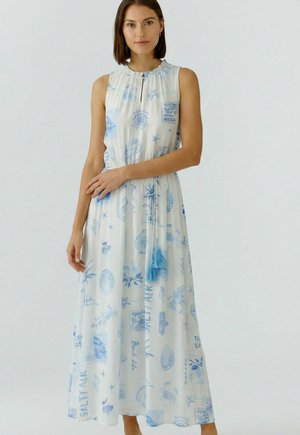 Frau trägt ein ärmelloses weißes Maxikleid mit hellblauen Muschel- und Ozeanmotiven und steht vor einem schlichten hellen Hintergrund.