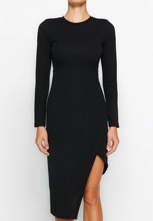 Vrouw draagt een zwarte jurk met lange mouwen, een hoge halslijn en een asymmetrische zoom die één been onthult.