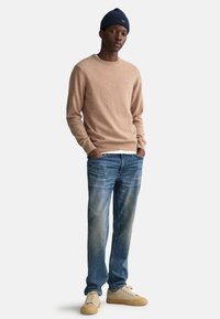 Beige gestrickter Pullover, blaue Jeans mit einem verwitterten Design und braune Sneakers. Trägt dazu eine marineblaue Mütze. Das Model steht mit den Händen in den Taschen.