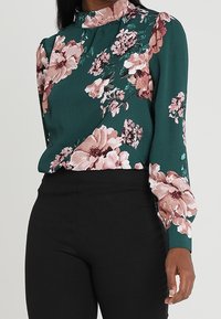 Blusa floreale verde con maniche lunghe, caratterizzata da fiori rosa e bianchi. Collo alto con una finitura testurizzata. Abbinata a pantaloni neri.