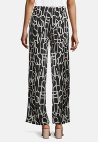 Osoba v černých širokých kalhotách se vzorem bílých abstraktních písmen, doplněná béžovými sandály na blokovém podpatku, zobrazená ze zadní strany.
