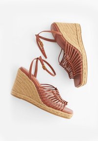 Sandales compensées en cuir marron avec talons plateforme en paille tissée et brides de cheville avec boucles dorées, disposées sur fond blanc.