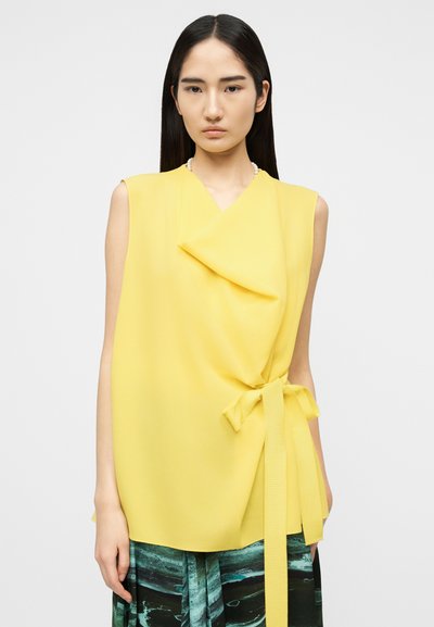 Femme portant un haut jaune sans manches noué à la taille et une jupe à motif vert, se tenant devant un fond clair uni.