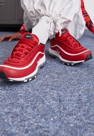 Osoba nosząca czerwono-białe buty sportowe Nike z czarnymi podeszwami, białe skarpetki i białe spodnie, siedząca na niebieskim wykładzinowym podłodze.