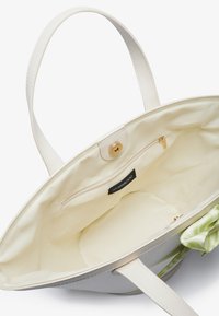 Tote taske med glat hvid læderoverflade, beige foring, indvendig lynlås lomme og et grønt mønstret stofaccitem i hjørnet.