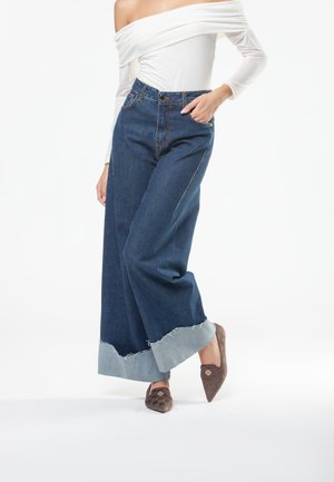 Wijduitlopende donkerblauwe spijkerbroek met versleten lichtblauwe boorden, gecombineerd met een strakke off-the-shoulder top en bruine puntige instappers.