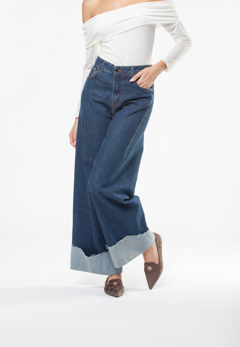 Jeans in denim blu scuro a gamba larga con orli sfilacciati blu chiaro, abbinati a un top attillato off-the-shoulder e mocassini marroni a punta.