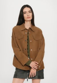 Giacca in suede marrone con colletto, due tasche al petto e chiusura con bottoni a pressione. Indossata sopra un top verde e una mini gonna nera.