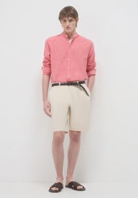 Camicia button-up rossa in lino con maniche arrotolate, pantaloni corti beigi su misura, cintura intrecciata marrone e sandali scuri a scivolo. Sfondo bianco uniforme.