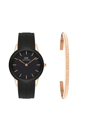 Daniel Wellington pulkstenim ir melns silikona josta, apaļš melns ciparnīca, zelta krāsas stundu rādītāji un slaids zelta akcentēts rokassprādze.