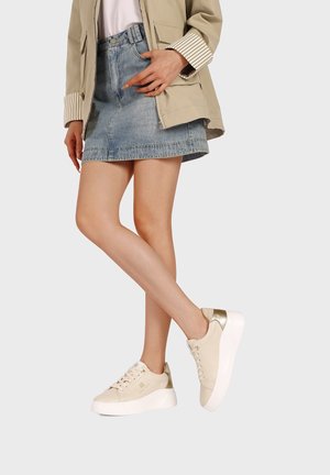 Donna con giacca beige, camicia bianca, gonna di jeans e sneakers beige con suole bianche, in piedi con le gambe incrociate su sfondo bianco.