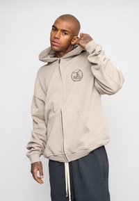 Sudadera beige con cremallera hecha de una tela suave, con un bolsillo delantero, puños acanalados y un logo en el pecho. Combinada con pantalones de chándal grises.