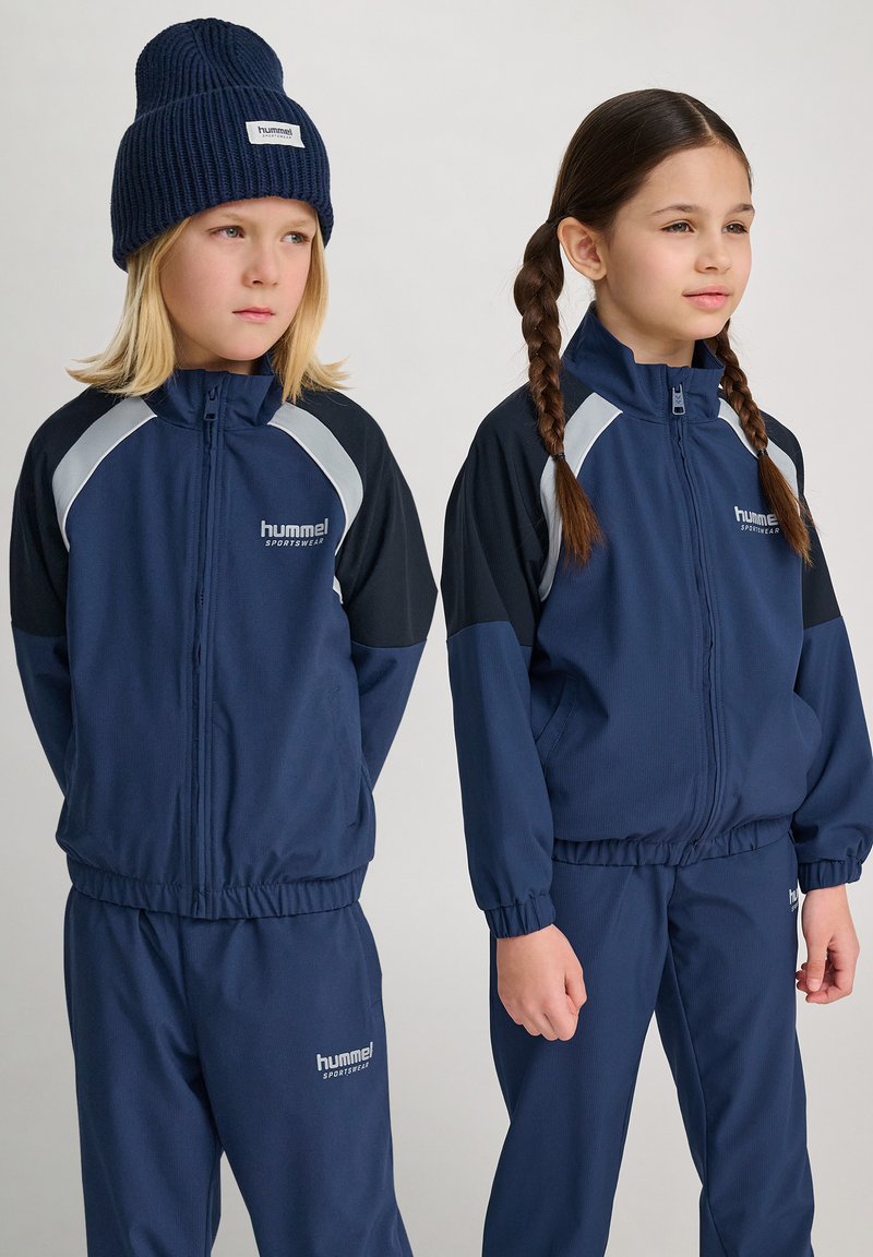 Marinblå sportjackor och byxor med grå detaljer, zippered front och logotyp. Ett barn bär en ribbad mössa; det andra har flätat hår.