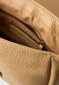 Beige strukturiertes Lederhandtascheninnere mit einem hellbraunen Stofffutter, das ein Reißverschlussfach und ein sichtbares Markenetikett sowie Nähdetails aufweist.