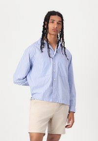 Camicia button-up a righe blu e bianche con una vestibilità rilassata, dotata di due tasche frontali e colletto, indossata con pantaloni beige.